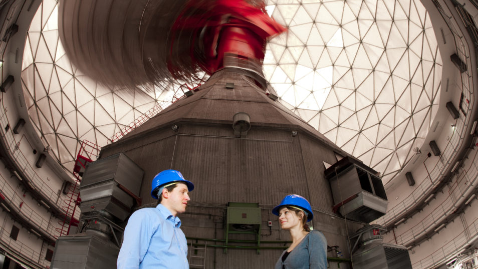 Ein Mann und eine Frau mit blauen Arbeitshelmen stehen in einem Gebäude mit Kuppeldach.